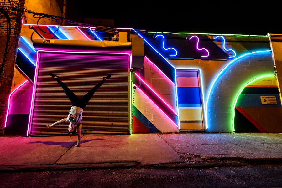 A woman doing a one handed handstand in south philadelphia in front of a neon light graffiti wall.